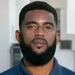 Headshot of REMAX Belize real estate agent Dyron Cadle, a man with a beard, against a blurred kitchen background. Belize REMAX Property Center